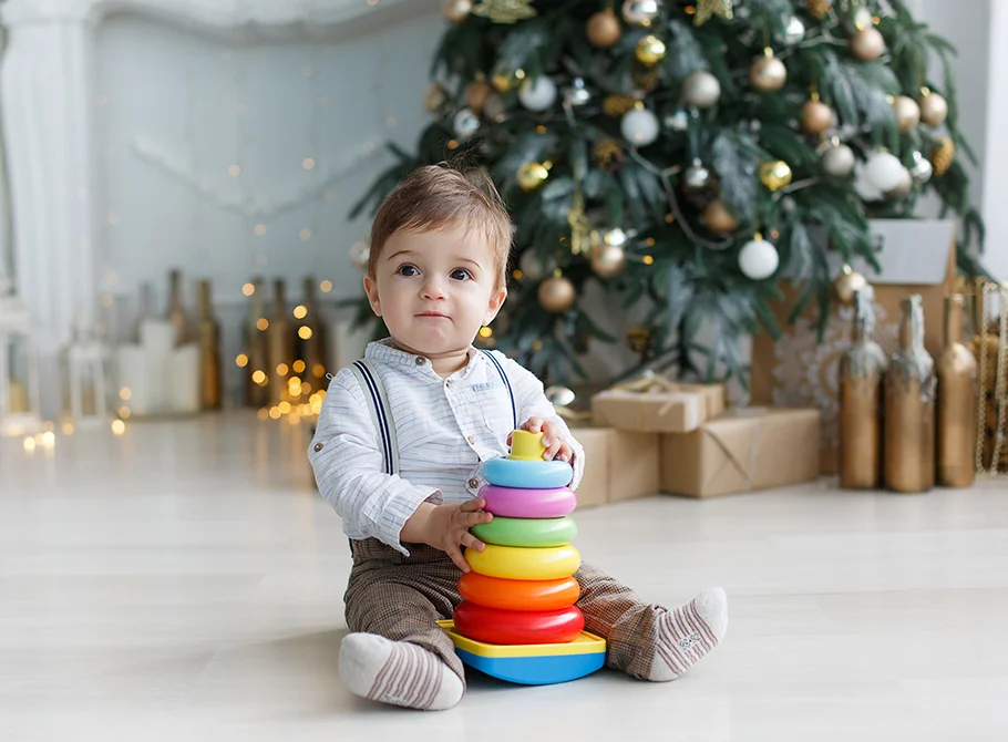 a cute kid in jumpsuit playing with stacking toy with Christmas tree and decorations in backgroung